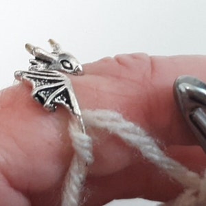 May include: A silver dragon ring with wings and a detailed design. The ring is being used as a stitch marker for crocheting.