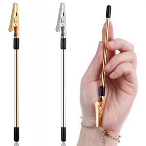 May include: A gold-colored and silver-colored alligator clip with a black tip and a long, thin, metal rod. The clip is attached to a bracelet. The clip is designed to hold items.