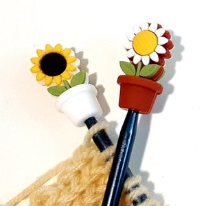 May include: Two knitting needles with flower-shaped stitch markers. One marker is a white daisy with a yellow center and green leaves. The other marker is a yellow sunflower with a black center and green leaves. Both markers are in pots.