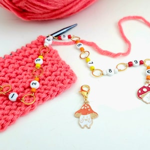 May include: A set of colourful mushroom stitch markers with numbers 1 through 9. The markers are attached to a gold chain with white beads. The markers are on a pink knitted fabric.