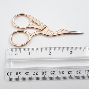 May include: Gold-coloured, decorative scissors with a bird-shaped handle. The scissors are approximately 7.5 cm long and have a pointed tip.