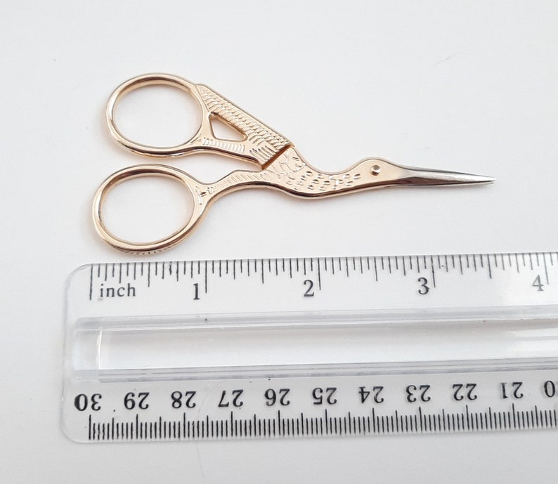May include: Gold-coloured, decorative scissors with a bird-shaped handle. The scissors are approximately 7.5 cm long and have a pointed tip.