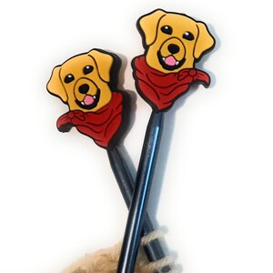 Puede incluir: Dos agujas de tejer con adornos de cabeza de perro amarillo y marr&oacute;n que llevan bandanas rojas. Las agujas son negras y azules.