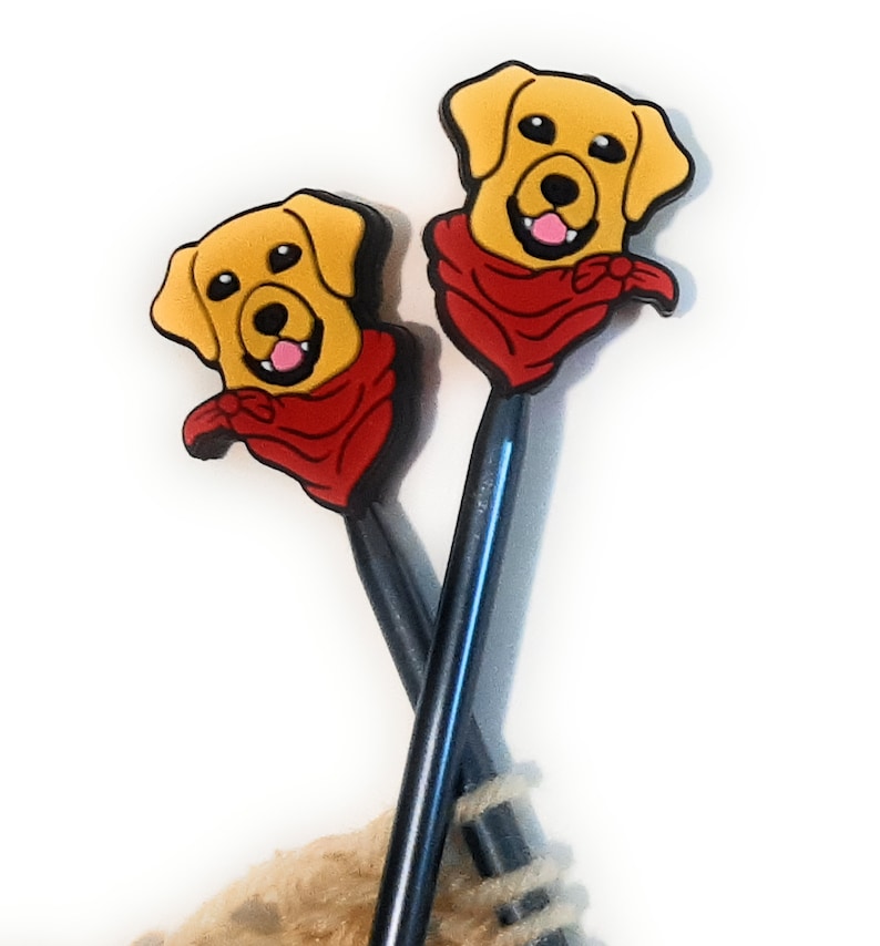 Puede incluir: Dos agujas de tejer con adornos de cabeza de perro amarillo y marr&oacute;n que llevan bandanas rojas. Las agujas son negras y azules.