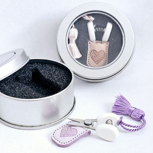 May include: A silver tin with a lid, containing a small leather pouch with a heart design and two white tools. A pair of small scissors with a lavender tassel and a matching leather heart are also present. The items are for crafting.
