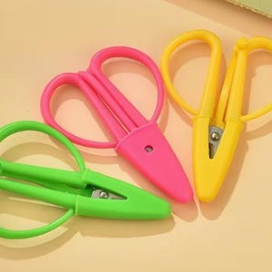 May include: Three pairs of colorful scissors in green, pink, and yellow. The scissors have rounded handles and pointed blades. The scissors are arranged on a light orange surface.