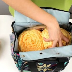 May include: A black and blue tote bag with floral print, featuring a zippered pocket and two handles. The bag is open and filled with yellow yarn balls.