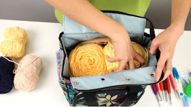 May include: A black and blue tote bag with floral print, featuring a zippered pocket and two handles. The bag is open and filled with yellow yarn balls.