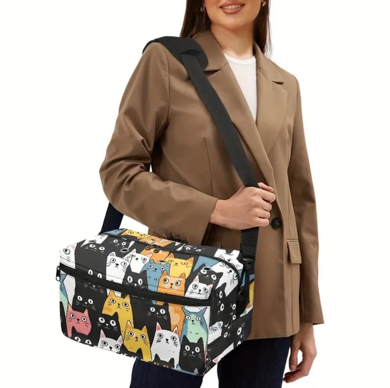 May include: A rectangular lunch bag featuring a repeating pattern of cartoon cats in various colors including black, white, orange, blue, and pink on a black background. The bag has a black adjustable shoulder strap and a black zipper closure.