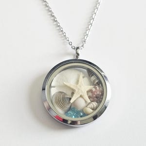 May include: A silver-toned necklace with a round, clear locket pendant. The pendant contains a starfish, seashells, and blue beads. The necklace is displayed against a white background, highlighting the beach-themed jewelry.