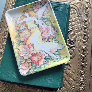 Può includere: Un piatto decorativo rettangolare con sfondo giallo, con un'illustrazione di fata circondata da rose. Il piatto è appoggiato su un libro verde, con una superficie dorata e ornata sullo sfondo. La parola "Summer" è visibile sul piatto.