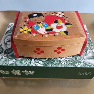 May include: A small wooden jewellery box with a painted design on the lid. The design includes a figure, flowers, and red details. The box is resting on a patterned box.