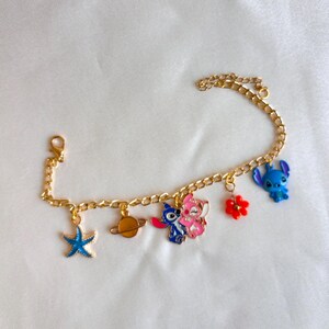 May include: Gold-colored charm bracelet featuring various colorful charms. Charms include a blue starfish, a planet, two cartoon characters, a red flower, and a blue cartoon character. The bracelet has a lobster clasp.
