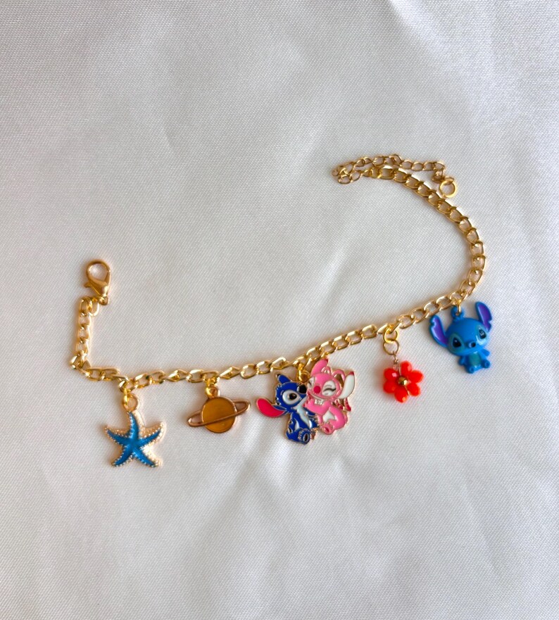 May include: Gold-colored charm bracelet featuring various colorful charms. Charms include a blue starfish, a planet, two cartoon characters, a red flower, and a blue cartoon character. The bracelet has a lobster clasp.