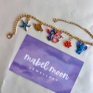 May include: Gold-colored chain bracelet with colorful enamel charms. Charms include a blue starfish, a planet, two Stitch characters, and a red flower. A purple card with the text "mabel moon JEWELLERY" is also present.