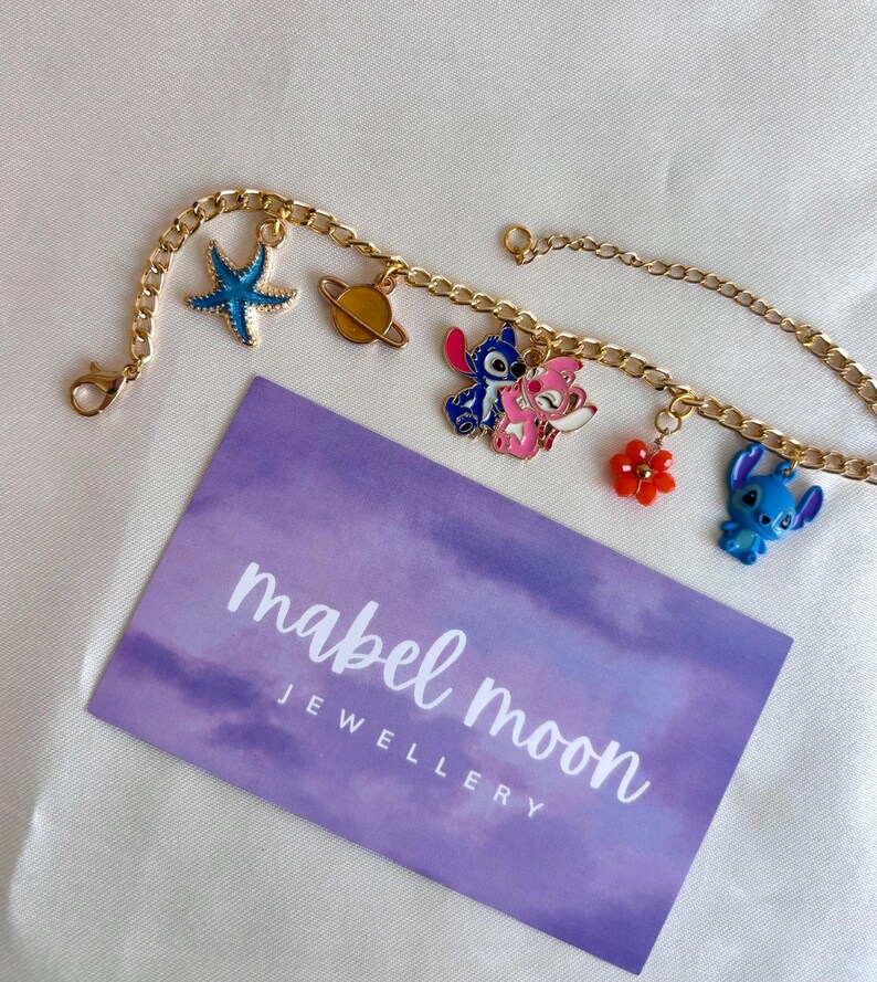 May include: Gold-colored chain bracelet with colorful enamel charms. Charms include a blue starfish, a planet, two Stitch characters, and a red flower. A purple card with the text "mabel moon JEWELLERY" is also present.