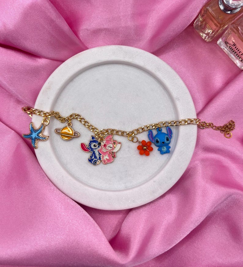 May include: Gold-colored charm bracelet featuring various enamel charms. Charms include a blue starfish, a planet, two cartoon characters, a red flower, and a blue cartoon character. The bracelet is displayed on a white circular tray against a pink background.