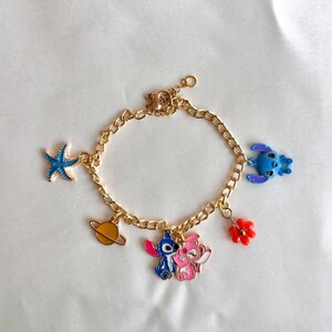 May include: Gold-colored charm bracelet featuring colorful enamel charms. Charms include a blue starfish, a yellow planet, a blue and pink cartoon couple, a red flower, and a blue cartoon character.