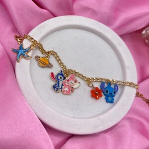 May include: A gold-colored chain bracelet with various charms, including a blue starfish, a yellow planet, and cartoon characters in blue, pink, and red. The bracelet is displayed on a white, circular tray against a pink satin background.