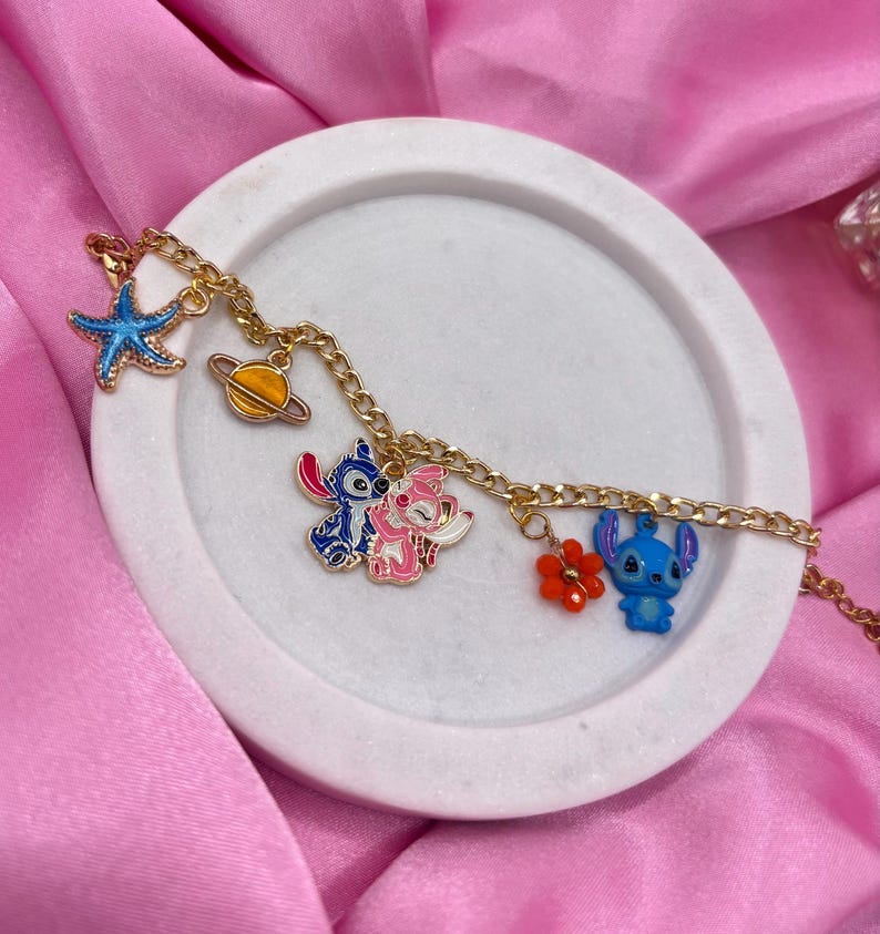May include: A gold-colored chain bracelet with various charms, including a blue starfish, a yellow planet, and cartoon characters in blue, pink, and red. The bracelet is displayed on a white, circular tray against a pink satin background.