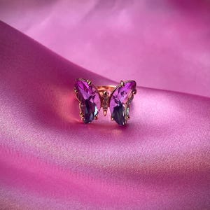 May include: A gold-colored ring featuring a butterfly design. The butterfly's wings are made of purple and blue-toned crystals. The ring is set against a pink satin background, creating a luxurious and eye-catching display.