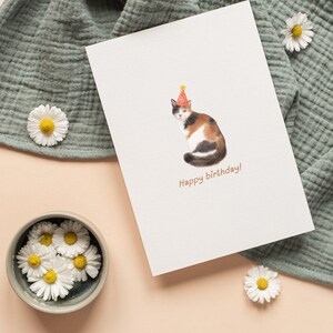 Puede incluir: Una tarjeta blanca con una ilustraci&oacute;n de acuarela de un gato calico con un sombrero de fiesta. El texto "Happy birthday!" est&aacute; impreso debajo del gato.