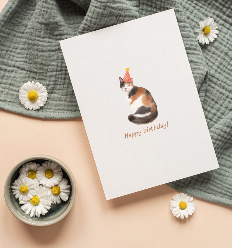 Puede incluir: Una tarjeta blanca con una ilustraci&oacute;n de acuarela de un gato calico con un sombrero de fiesta. El texto "Happy birthday!" est&aacute; impreso debajo del gato.