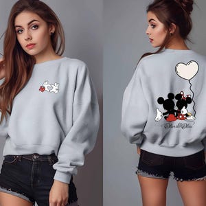May include: Light gray sweatshirt featuring a cartoon design on the front and back. The front has a white rabbit forming a heart with its hands and a red flower. The back has Mickey and Minnie Mouse with a heart balloon.