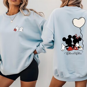 May include: Light blue sweatshirt featuring a cartoon rabbit design with a heart and flowers on the front. The back of the sweatshirt has a Mickey and Minnie Mouse graphic with a heart balloon. The text "Oliver & Olivia" is below the graphic.
