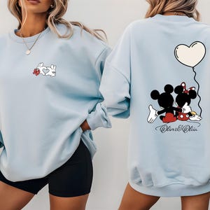 May include: Light blue sweatshirt featuring a cartoon rabbit design with a heart on the front. The back of the sweatshirt has a Mickey and Minnie Mouse graphic with a heart balloon. The text "Oliver & Olivia" is below the graphic.