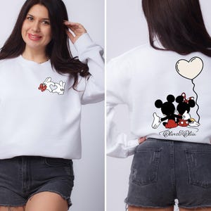 May include: White sweatshirt featuring Mickey and Minnie Mouse graphics. The front displays Mickey's hands forming a heart with a red bow. The back shows Mickey and Minnie with a heart balloon. The text "Oliver & Olivia" is below.