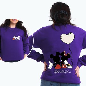 May include: A purple sweatshirt featuring a Mickey and Minnie Mouse design. The back of the sweatshirt shows the couple embracing under a heart-shaped balloon. The front has a hand-heart design with a red flower.