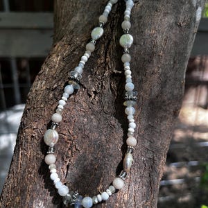 May include: A white and gray beaded necklace with silver accents. The necklace is made with various shapes and sizes of beads, including round, oval, and irregular shapes. The necklace is hanging on a tree branch.