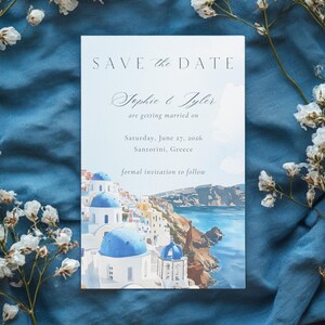 May include: A rectangular "Save the Date" card with a watercolour illustration of Santorini, Greece. The card features blue-domed buildings, the sea, and the text "SAVE the DATE" and "Sophie & Tyler are getting married on Saturday, June 27, 2026, Santorini, Greece."