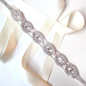 Puede incluir: Un cinturón delicado de pedrería plateada con un patrón repetitivo de óvalos entrelazados. El cinturón se muestra sobre una cinta de satén crema.
