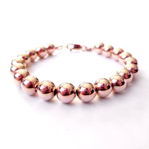 May include: A rose gold beaded bracelet with a shiny finish. The bracelet is made of round beads and has a lobster clasp closure.