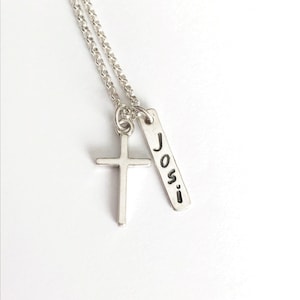 May include: A silver-colored necklace featuring a cross pendant and a rectangular tag. The tag is engraved with the name "Josi". The necklace has a delicate chain and is set against a white background.