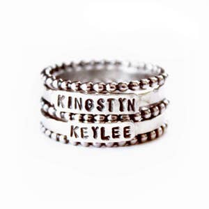 Peut inclure: Bague en argent avec quatre bandes, chacune avec un bord perlé. La bande supérieure porte le nom "KINGSTYN" gravé, et la deuxième bande porte le nom "KEYLEE" gravé.