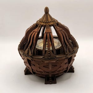 May include: A detailed, brown decorative compass in a cage-like structure. The compass is visible through the openwork design of the cage. The base has a decorative pattern, and the top has a pointed finial.