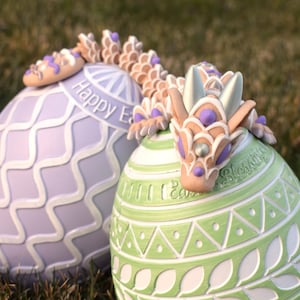 May include: Two decorated Easter eggs, one purple and white with the text "Happy Easter" and one green and white with the text "Easter Blessings", with a small, light brown dragon figurine with purple accents sitting on top of the green egg.