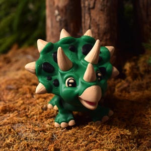 May include: A green and brown toy dinosaur with a large horn and frill. The dinosaur has large, black eyes and a friendly expression.