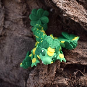 May include: A green and yellow 3D printed dragon figurine with a clover design. The dragon is perched on a tree trunk.