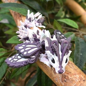 May include: A white and purple dragon figurine with butterfly wings, perched on a branch. The dragon has a detailed, intricate design and a whimsical, fairy-like appearance.