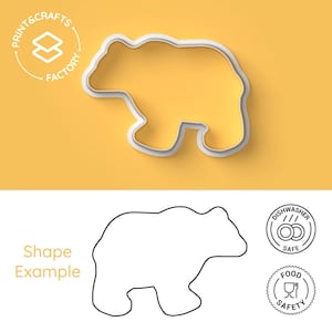 Op de afbeelding: Een witte koekjesvorm in de vorm van een beer met het logo Print & Crafts Factory. Hieronder toont een zwarte omtrek de beervorm, vergezeld van twee ronde iconen met de tekst Dishwasher Safe en Food Safety, die de productkenmerken aangeven.