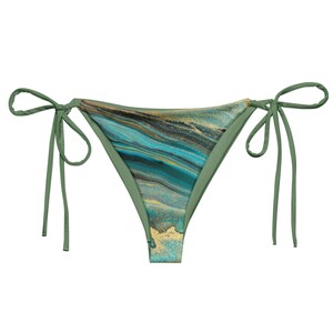 May include: A bikini bottom with a unique abstract design. The front panel features a swirling pattern of teal, gold, and black, while the edges and ties are a solid olive green. The ties are long and can be adjusted for a comfortable fit.