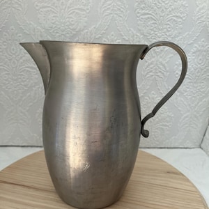 May include: A silver pitcher with a rounded body and a curved handle. The pitcher has a smooth, shiny surface.