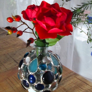 May include: A decorative glass vase with a mosaic design, filled with vibrant red roses and berries. The vase features a variety of blue, white, and clear glass pieces, creating a unique and artistic floral arrangement. The vase sits on a wooden surface.