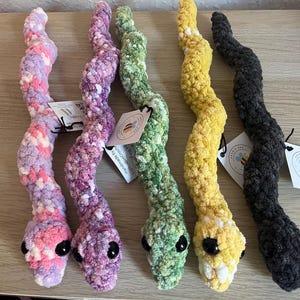May include: Five plush snakes made of soft, textured yarn in various colours including pink and purple variegated, solid green, yellow, and dark grey. Each snake features black plastic eyes and a small white paper tag attached to the body.