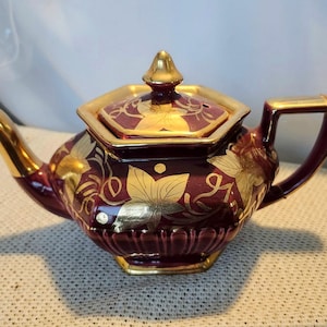 May include: A burgundy and gold teapot with a lid. The teapot features gold leaf designs and trim. The handle and spout are also gold. The teapot is sitting on a textured surface.