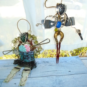 May include: Two handmade ornaments made from recycled materials. The ornament on the left is a teal blue and green color with a pink and blue candy cane. The ornament on the right is a silver and gold color with a blue bead and red legs.